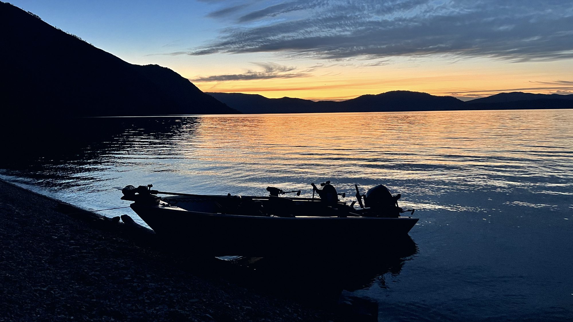 A boat pulled up on a gravel beach at first light with mountains behind.