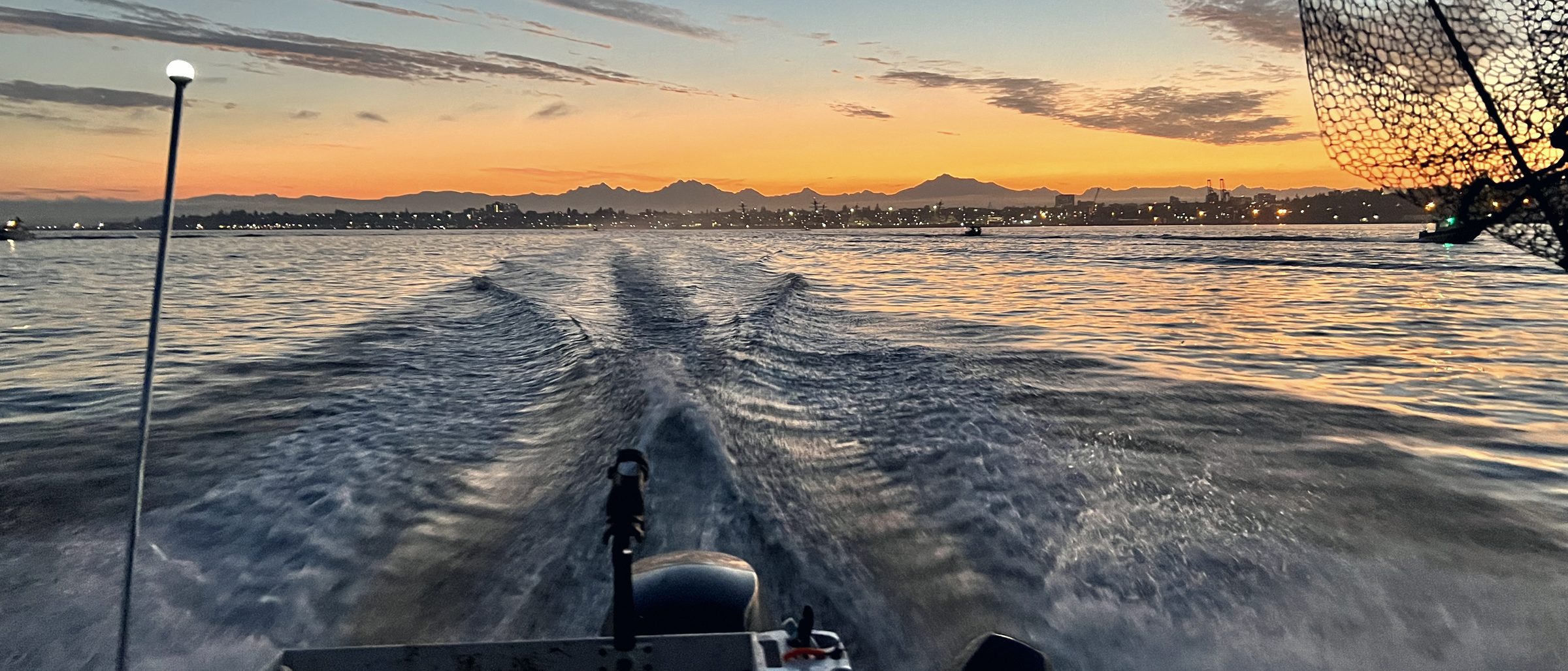 Wake trailing behind a boat running out before daylight, landing lights on the shoreline behind.